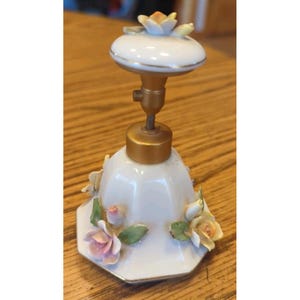 May include: A white porcelain bell featuring a gold-colored metal handle and a top knob adorned with a sculpted flower. The bell body is decorated with three-dimensional ceramic roses in shades of pink and yellow with green leaves.