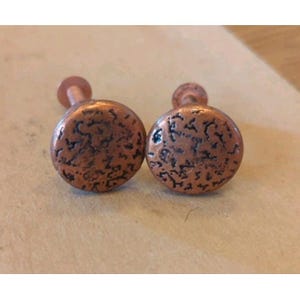 May include: Two circular copper-toned metal knobs featuring a textured, hammered surface with dark, recessed detailing. Each knob has a smooth, rounded face and a short, cylindrical stem extending from the back for mounting purposes.