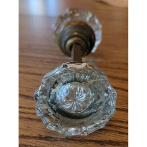 May include: A clear, faceted glass doorknob with a brass-colored base and a dark metal rod. The knob has a starburst pattern in the center and a decorative, vintage design. The doorknob is likely for a cabinet or door.