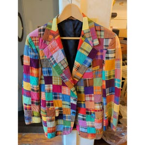 May include: A blazer constructed from a patchwork of various plaid and solid-colored fabric squares in shades of red, blue, yellow, green, orange, and purple. The garment features a notched lapel, two front buttons, and a solid black interior lining.