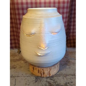 May include: A cream-colored ceramic vessel shaped like a human head with closed eyes, a nose, and a mouth. The piece features cutouts at the eyes and mouth that emit a warm light from within. It is mounted on a cylindrical wooden base.