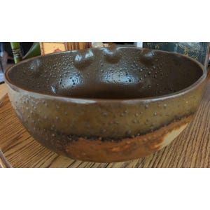 May include: A handcrafted, brown ceramic bowl with a textured surface and a unique, organic shape. The bowl features a darker brown interior and a lighter brown band around the exterior, adding visual interest. This decorative bowl is perfect for serving or display.
