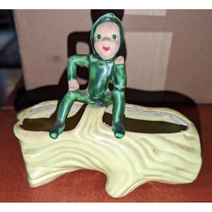 May include: A ceramic planter shaped like a light yellow log with a textured bark surface. A small, green-clad figure with a hood, painted facial features, and a red mouth sits perched on top of the log, which features a hollow opening.
