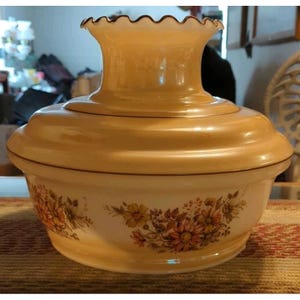 May include: A round, cream-colored glass serving dish with a matching lid featuring a ruffled top edge. The exterior of the bowl is decorated with a floral pattern in shades of orange, yellow, and brown, showcasing a classic, rounded silhouette.