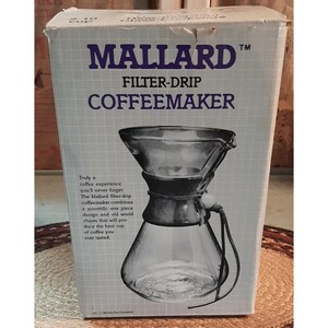May include: A Mallard filter-drip coffeemaker in its original box. The box is white with blue text that reads "MALLARD FILTER-DRIP COFFEEMAKER". The coffeemaker is glass and has a unique design. The box also includes text about the coffee experience.