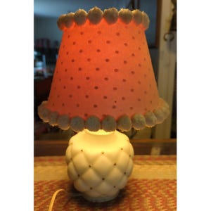 May include: A vintage table lamp with a white, quilted base and a coral-colored lampshade. The lampshade is decorated with dark polka dots and trimmed with white pom-poms. The lamp is turned on, emitting a warm glow.
