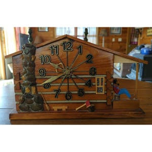 May include: A wooden cabin-shaped clock featuring a stone chimney on the left, a porch on the right with a seated figure, and a central clock face with black numbers. A small wooden bench and an axe are placed at the base of the cabin structure.