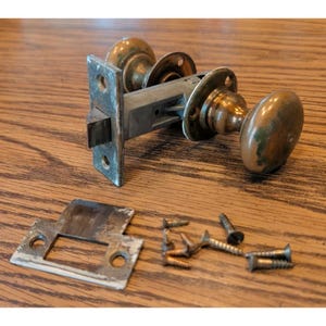 May include: A vintage brass door knob set with a rectangular latch plate, a separate metal strike plate, and several small metal screws. The brass components show signs of age and wear, featuring a rounded knob design and a metallic latch mechanism.