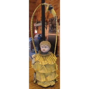 May include: A vintage doll with a pale face and painted features, wearing a multi-layered yellow ruffled dress with a dark collar. The doll is attached to a tall, arched yellow handle that extends upwards, creating a basket-like structure.
