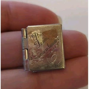 May include: A small, silver-colored book charm with a hinged spine. The cover is engraved with the words "The Teen Commandments" in an elegant script. The charm is held between fingers.