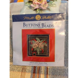 May include: A Mill Hill Buttons & Beads Winter Series craft kit in a clear plastic package. The label features a cross-stitch design of pine branches, red ribbons, and pinecones on a dark background. The design size is 5.25 inches by 5.25 inches.