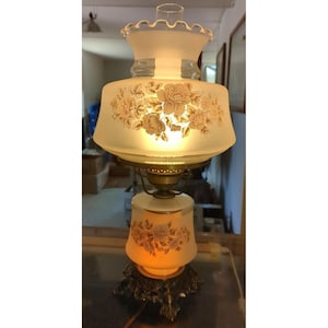 May include: A vintage-style table lamp with a frosted glass shade and base, adorned with floral patterns in shades of beige and brown. The lamp features a brass-colored metal base and a clear glass chimney. The lamp is lit, emitting a warm glow.