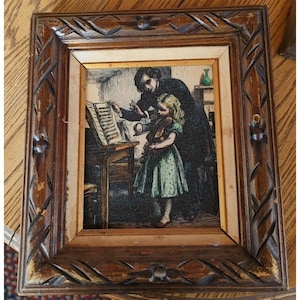 May include: A rectangular wooden picture frame with carved leaf patterns surrounding a print of a person teaching a child to play the violin. The child wears a light green dress, while the adult stands behind them near a music stand.