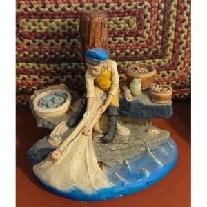 May include: A figurine of a fisherman wearing a blue cap, yellow shirt, and black boots, sitting on a rock while mending a beige fishing net. The scene includes two small wooden buckets, a jug, and a blue base representing water waves.