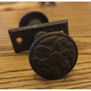May include: A dark bronze-toned metal door knob featuring a circular face with an embossed floral pattern. The knob is attached to a rectangular backplate that includes two circular screw holes on either side of the central stem mechanism.