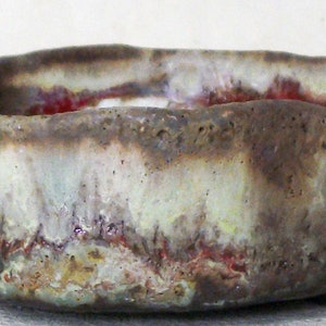 Puede incluir: Un pequeño cuenco redondo de cerámica con una superficie texturizada y desigual. El cuenco está esmaltado en tonos marrones, blancos y rojos, creando un efecto moteado.