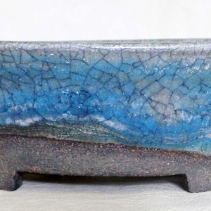 May include: A rectangular bonsai pot with a textured, crackled glaze. The upper section is a vibrant blue, transitioning to a darker, earthy brown base. The pot has a low profile with small feet.