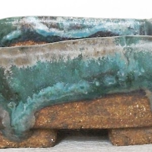 Puede incluir: Una maceta de bonsái rectangular de cerámica con un esmalte verde, azul y marrón. La maceta tiene tres patas y una abertura rectangular.