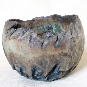 May include: A ceramic bowl with a rough, textured surface and a mottled brown, green, and gray glaze. The bowl has a wide, rounded rim and a deep, rounded interior.