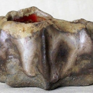 May include: A small, irregularly shaped ceramic planter with a rough, textured surface. The planter is a mottled brown, gray, and white color with a red glaze on the inside.