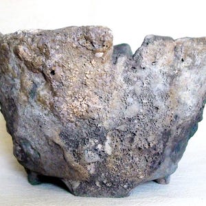 Puede incluir: Un cuenco de cerámica texturizado, con un aspecto rugoso y envejecido. El cuenco presenta una paleta de colores gris y marrón con una superficie porosa y pequeños pies. El diseño único sugiere una estética rústica o artesanal.