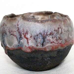May include: A small, round ceramic bowl with a dark brown glaze and a white and red glaze on the top. The bowl has a unique, textured surface.