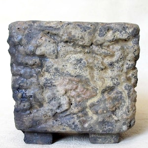 Peut inclure: Un pot carré texturé avec un aspect rugueux semblable à de la pierre. Le pot est principalement gris avec des touches de beige et de marron, et repose sur quatre petits pieds rectangulaires. La surface a une texture rugueuse et inégale.