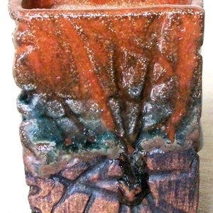 May include: A square, terracotta-coloured ceramic planter with a rough, textured surface. The planter has a mottled glaze with shades of orange, brown, and green.