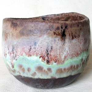 Peut inclure: Un bol en céramique fait à la main, de forme organique unique. Le bol présente un mélange de tons terreux, notamment marron, vert et crème, avec une surface texturée. L'intérieur est brun foncé et l'extérieur a une finition glacée mouchetée.