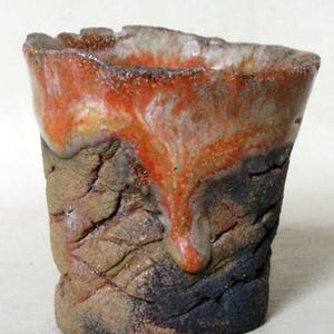 Peut inclure: Une petite tasse en céramique faite à la main avec une surface rugueuse et texturée. La tasse est émaillée dans des tons d'orange, de brun et de noir, avec un glaçage blanc en goutte qui coule sur le côté.