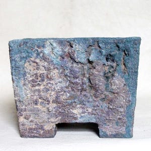 May include: A rectangular ceramic planter with a rough, weathered texture. The surface features a mottled color palette of deep teal, earthy brown, and muted gray tones. The base includes two short, sturdy feet with a central open space between them.
