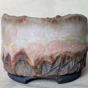 Puede incluir: Una maceta de bonsái de cerámica con una superficie texturizada y desigual. La maceta es de color marrón claro con vetas de color rosa, amarillo y blanco. La base de la maceta es de color marrón oscuro.