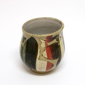 May include: A ceramic cup with a speckled beige base featuring bold, vertical brushstrokes in black, dark green, and red. The vessel has a rounded, bulbous body, a slightly flared rim, and a smooth, light-coloured interior finish.