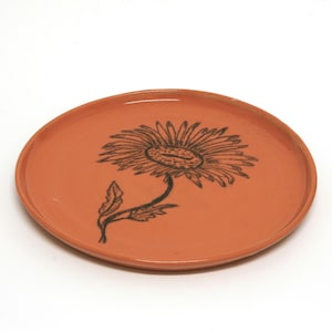May include: A terracotta-colored ceramic plate with a black floral design. The flower is stylized and has a large center with petals radiating outward.