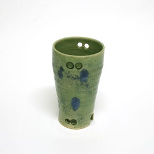 May include: A handmade green ceramic tumbler with a textured surface and blue speckles. The tumbler has two sets of two small holes near the top and bottom. The ceramic cup is a unique piece of pottery.