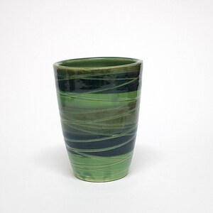 May include: A cylindrical ceramic tumbler featuring a glossy finish with alternating horizontal bands of dark forest green and lighter lime green, overlaid with thin, swirling, hand-painted white lines that wrap around the entire exterior surface.