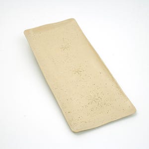 May include: A rectangular, beige ceramic serving platter with a speckled texture. The platter features three embossed snowflake designs. The edges are slightly irregular, giving it a handmade appearance. The background is plain white.