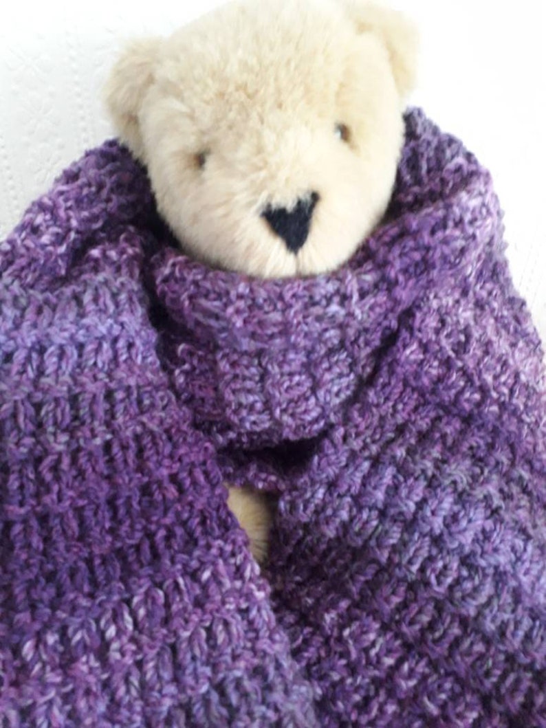 May include: A purple and white knitted scarf with a teddy bear wearing it. The scarf is soft and fluffy, and the bear is cute and cuddly.