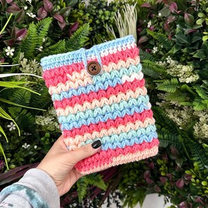 Peut inclure: Une housse de tablette crochetée à la main, aux couleurs vives. La housse présente des rayures horizontales roses, bleu clair, crème et blanches. Un bouton en bois ferme le rabat supérieur. La housse est tenue devant un fond de verdure.