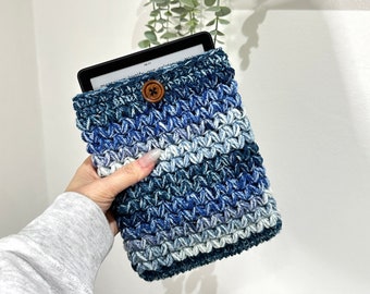 May include: A blue and white crocheted e-reader cover with a wooden button. The cover is made with a textured stitch and has a gradient of color from dark blue to light blue.