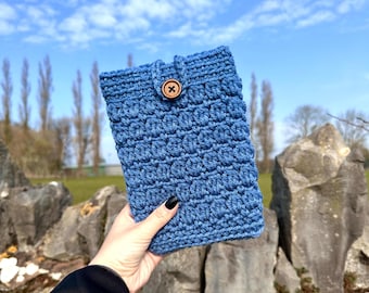 May include: A blue crocheted tablet sleeve with a textured pattern and a wooden button closure. The sleeve is held against a backdrop of a blue sky and a stone wall, showcasing the handmade craftsmanship.
