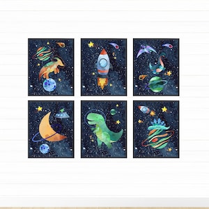以下が含まれることがあります： 宇宙をテーマにした水彩画の額装が6点。それぞれ異なる恐竜や宇宙関連のオブジェクトが描かれており、ロケット、UFO、三日月などがあります。プリントは、白い星が散りばめられた濃い青色の背景に配置されています。