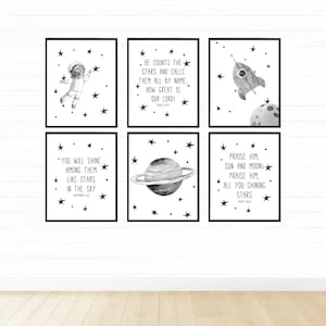 Peut inclure: Six impressions d'art encadrées sur le thème de l'espace en noir et blanc. Les impressions présentent un astronaute, une fusée, une planète et des citations inspirantes avec des illustrations d'étoiles. Les impressions sont affichées sur un mur blanc au-dessus d'un sol en bois.