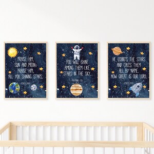 Op de afbeelding: Drie ingelijste kunstprints met een ruimtethema. Elke print heeft een donkerblauwe sterrenachtergrond, gouden sterren en aquarel illustraties van planeten, de zon en een raket. Tekst uit Psalmen en Filippenzen is inbegrepen.