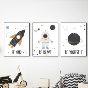 Puede incluir: Tres impresiones enmarcadas blancas con ilustraciones en negro y dorado de un cohete, un astronauta y un planeta. Las impresiones tienen las palabras "Sé amable", "Sé valiente" y "Sé tú mismo" en ellas.