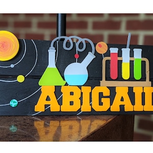 Puede incluir: Un letrero de madera con el nombre Abigail en letras naranjas. El letrero está decorado con un sol amarillo, planetas y vasos de precipitados científicos.