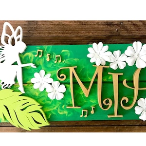 Könnte beinhalten: Ein Holzschild mit grünem Hintergrund und dem Namen "MIA" in goldenen Buchstaben. Das Schild ist mit einer weißen Fee, weißen Blumen und grünen Blättern verziert.