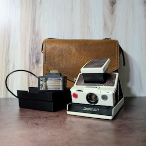 Puede incluir: Una cámara Polaroid SX-70 Land Camera Model 2 vintage, con un accesorio de flash negro y una funda de cuero marrón. La cámara es blanquecina con una parte superior negra y marrón, y un botón rojo.