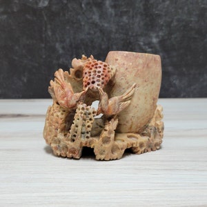 Puede incluir: Una escultura de piedra tallada a mano de dos pájaros posados en una rama, con un pequeño jarrón redondo en el centro. La escultura está hecha de una piedra marrón claro con acentos rojos.