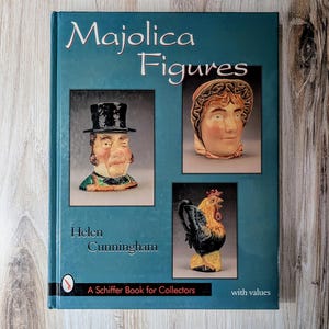 Puede incluir: Un libro verde azulado titulado "Majolica Figures" de Helen Cunningham, con imágenes de figuras de cerámica. La portada incluye un hombre con sombrero de copa, la cabeza de una mujer y un gallo. El libro está etiquetado como "A Schiffer Book for Collectors."
